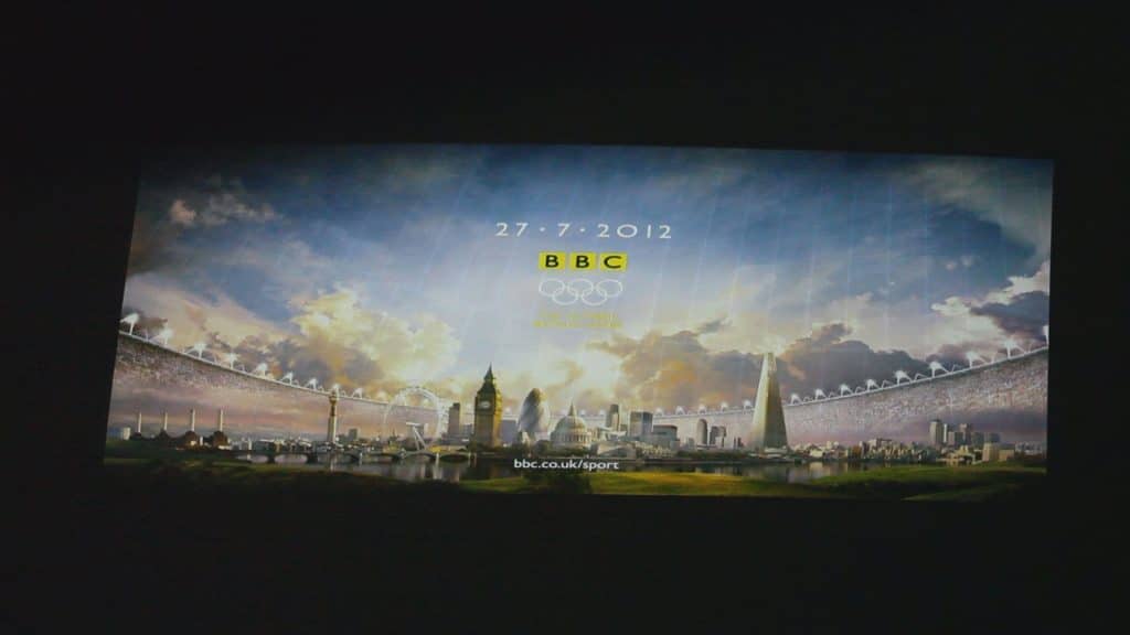 Slajd projekcji z festiwalu BlueBox 2013 w olsztyńskim Planetarium, przedstawiający londyńskie symbole i logo BBC, datę i wiadomość.