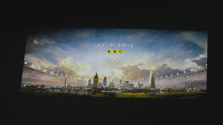 Slajd projekcji z festiwalu BlueBox 2013 w olsztyńskim Planetarium, przedstawiający londyńskie symbole i logo BBC, datę i wiadomość.
