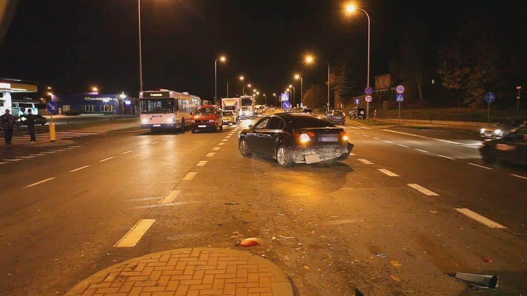 Uszkodzony samochód na środku ulicy Sielskiej po kolizji, w tle pojazdy i autobusy, nocna scena.