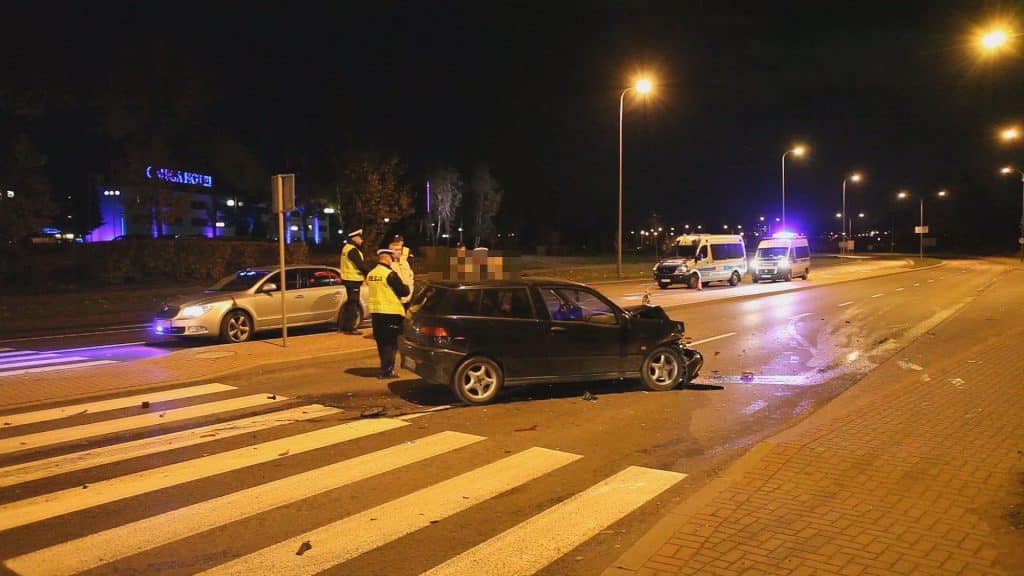 Zniszczony samochód na pasach po kolizji, w tle radiowozy i policjanci na ulicy nocą.