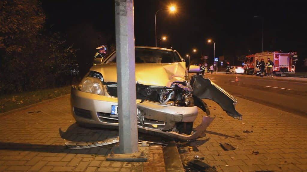 Uszkodzony samochód po kolizji na ulicy Sielskiej w Olsztynie, części pojazdu na chodniku, w tle służby ratunkowe i inni uczestnicy.