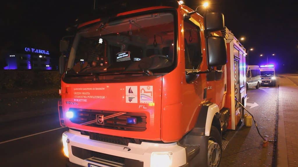 Czerwony wóz strażacki na ulicy o zmroku, w tle inne pojazdy i niebieskie światła syren policyjnych.