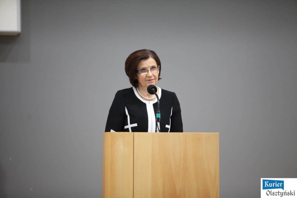 Kobieta przemawia z podium podczas uroczystości w urzędzie, w tle szara ściana.