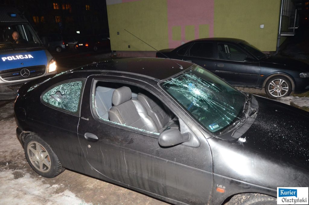 Uszkodzony czarny samochód z wybitymi szybami, na parkingu wieczorem, z policjantem w tle.