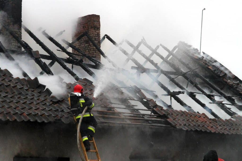 Strażak gasi pożar dachu spiżowskiego budynku podczas akcji ratowniczej, w tle widoczne zgliszcza i dym.