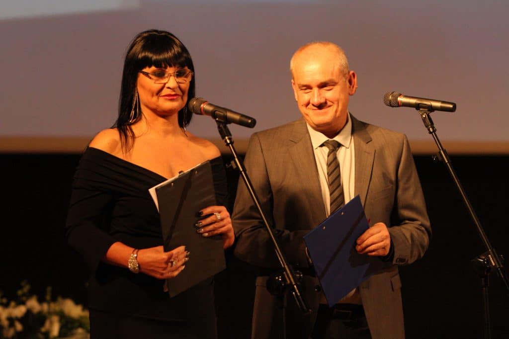 Dwóch uhonorowanych sportowców podczas gali w filharmonii, kobieta w czarnej sukni, mężczyzna w garniturze.