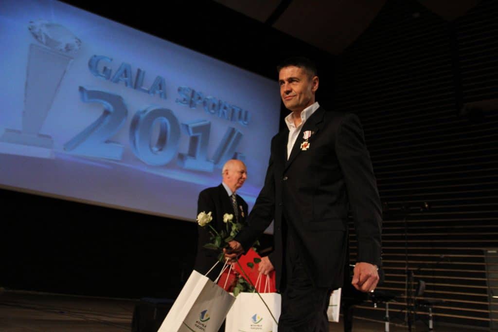 Mężczyzna w garniturze z odznaczeniami i kwiatami na scenie podczas gali sportu, w tle ekran z napisem "GALA SPORTU 2014".