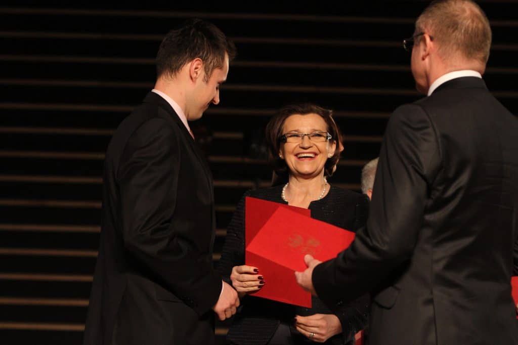 Osoba wręczająca medal sportowcowi podczas gali w filharmonii, kobieta uśmiecha się.