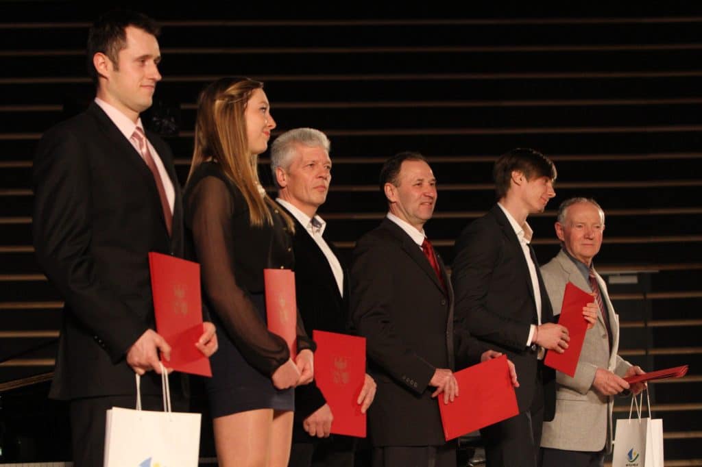 Grupa wyróżnionych sportowców na scenie podczas gali w filharmonii, trzymających czerwone dyplomy.