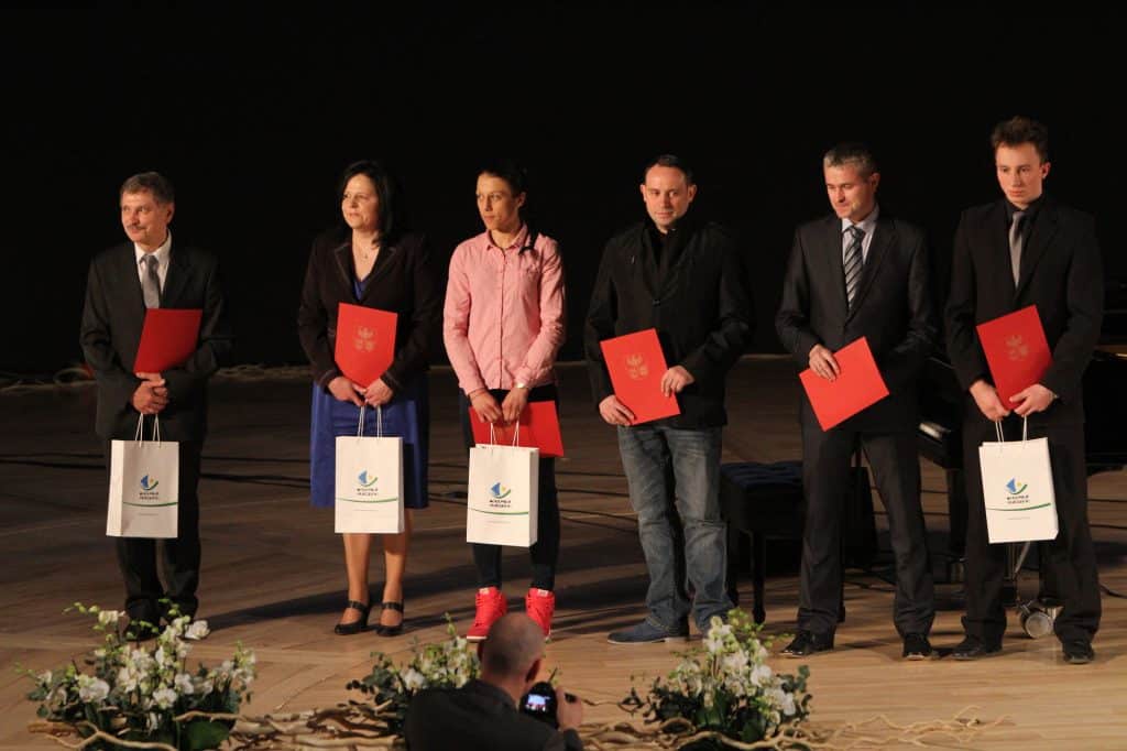 Siedem osób na scenie podczas uroczystości, trzymają czerwone foldery i torby z logo organizatora.