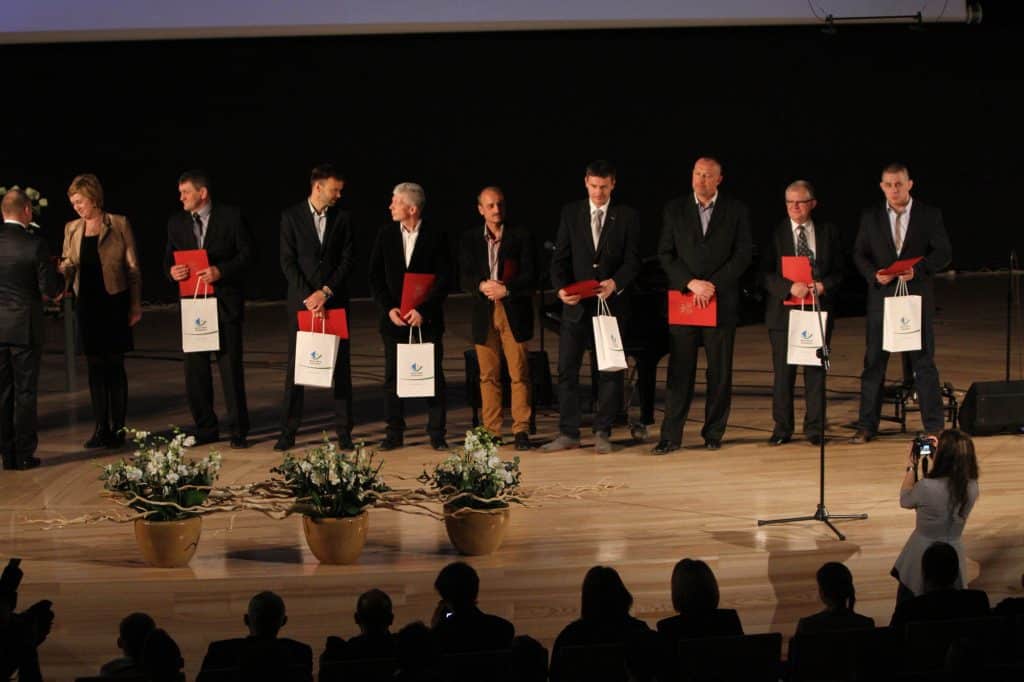Grupa sportowców na scenie podczas gali, trzymają odznaki i torby, w tle ciemne tło i kwiatowe ozdoby.