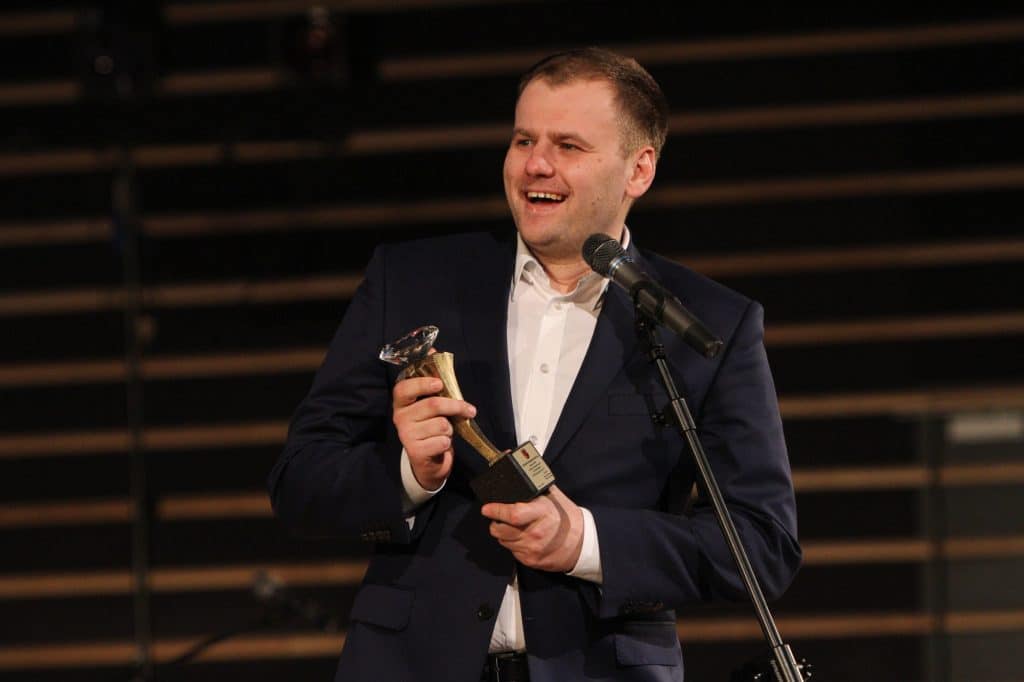 Mężczyzna w garniturze trzymający nagrodę podczas gali sportu w filharmonii.