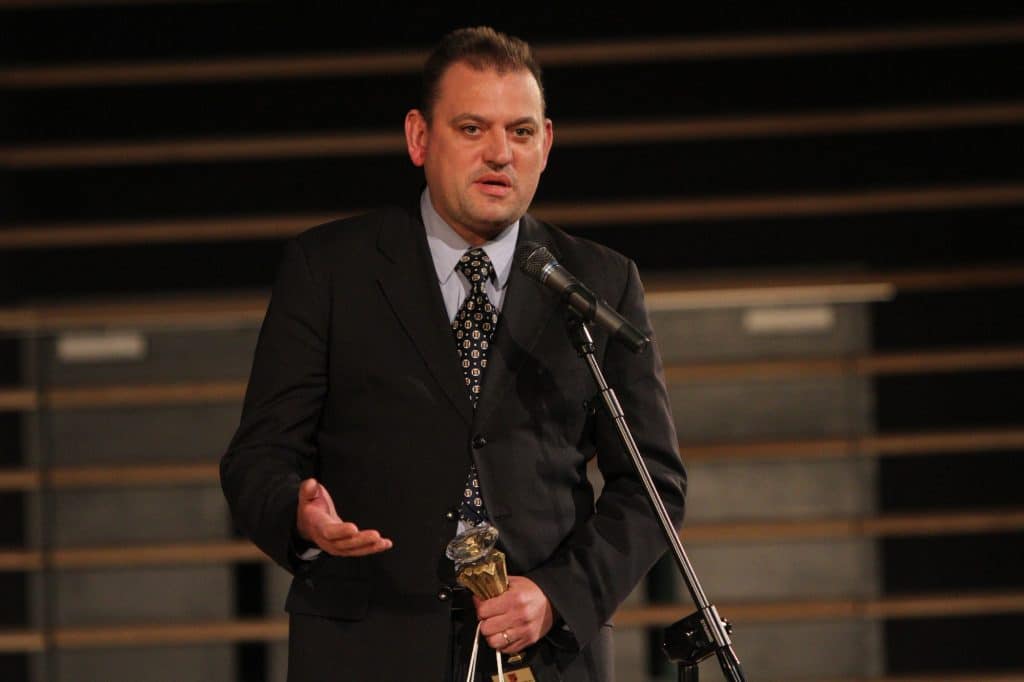 Mężczyzna w garniturze przemawia na scenie podczas gali sportowej, trzymając nagrodę. W tle pusta scena lub trybuny.