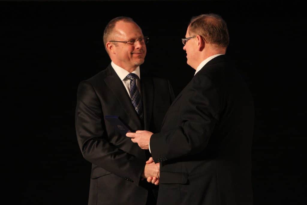 Dwie osoby w formalnych strojach podczas uroczystości na scenie, jedna z nich wręcza odznakę drugiej.