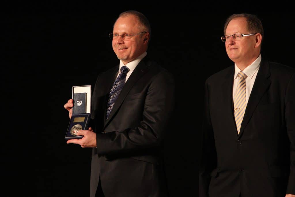 Dwóch mężczyzn w garniturach podczas ceremonii wręczenia odznaki na gali sportowej.
