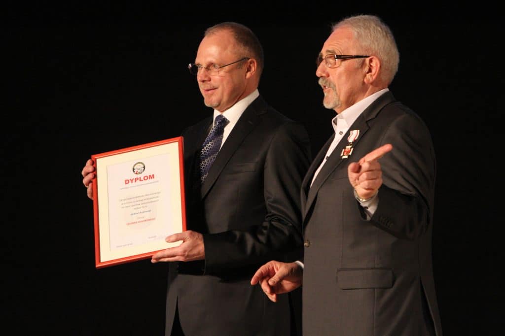 Dwóch mężczyzn na scenie podczas ceremonii wręczania odznaczeń, jeden trzyma dyplom, drugi wskazuje na coś. Obaj w garniturach.