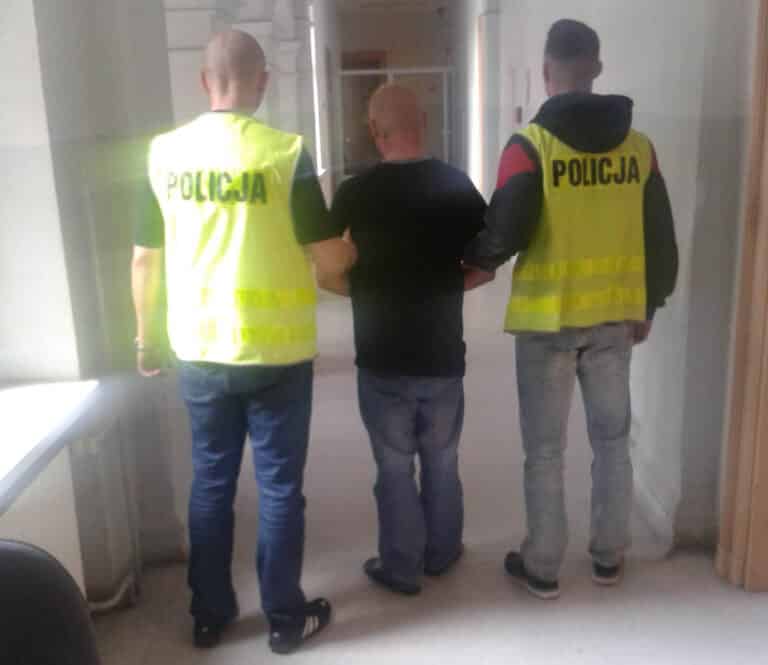 Mężczyzna w rękach policji, trzymany przez dwóch funkcjonariuszy, w pomieszczeniu.
