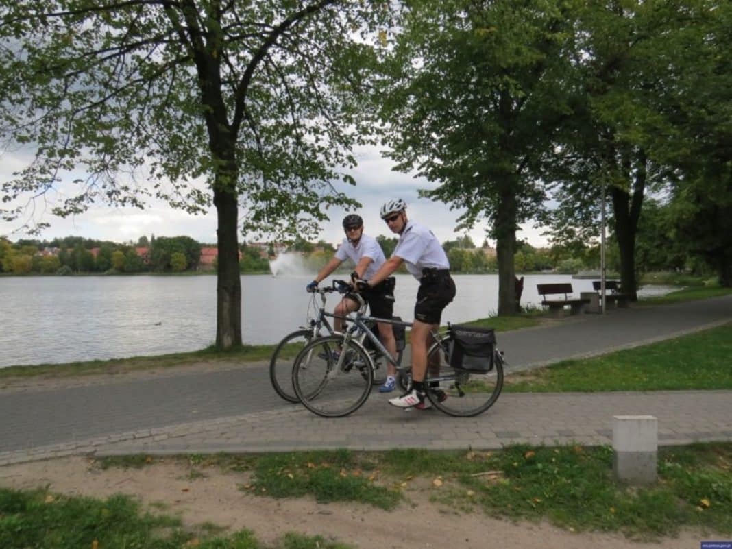 Dwóch policjantów na rowerach patrolowych rozmawia z mężczyzną nad jeziorem, który ma owiniętą rękę z krwią.