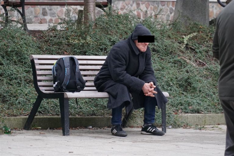 Bezdomny mężczyzna siedzi na ławce w parku, ubrany w ciemne ubrania, z plecakiem obok.