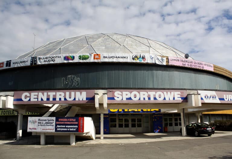 Widok na budynek Centrum Sportowego z dużym napisem na fasadzie, na dachu banery reklamowe i ogłoszenia.