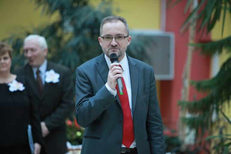 Mężczyzna w garniturze trzyma mikrofon podczas wydarzenia, w tle osoby w eleganckich strojach.