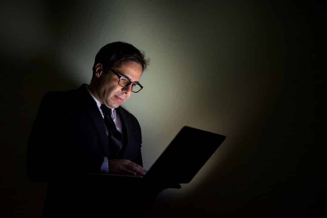 Mężczyzna w garniturze pracujący przy laptopie w słabym świetle.