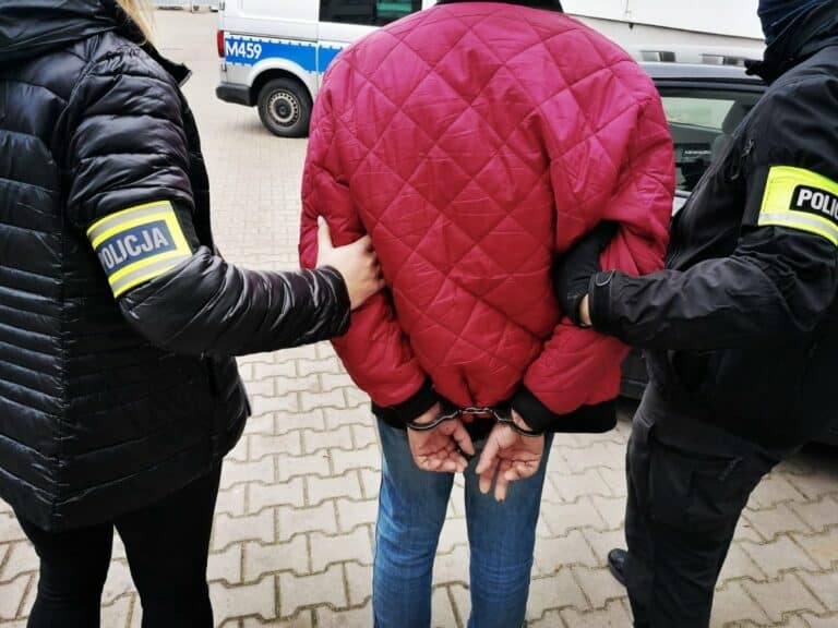 Policjanci zatrzymują mężczyznę z rękami zakutymi w kajdanki, w tle radiow&oacute;z policyjny.