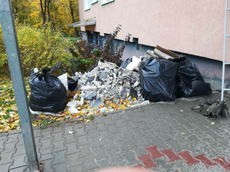 Odpady remontowe porozrzucane przy ścianie budynku na chodniku, w workach i na ziemi.