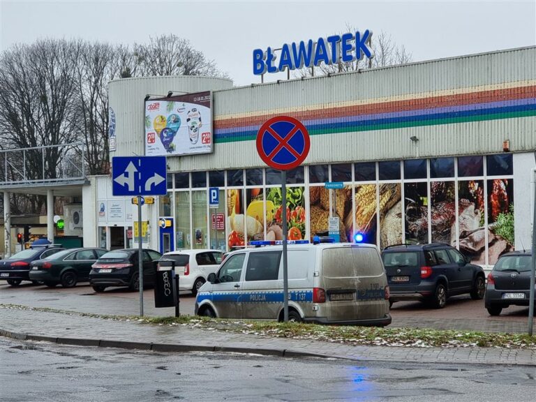 Policja na parkingu przy sklepie spożywczym w Olsztynie, w tle reklamy i budynek sklepu.