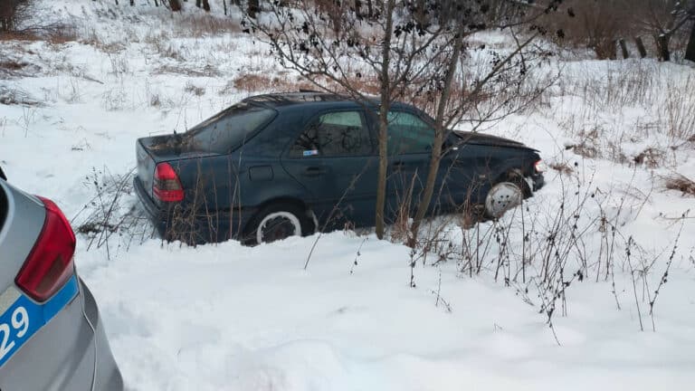 Uszkodzony czarny samoch&oacute;d na zaśnieżonej drodze, zniszczony po wypadku, w pobliżu patrol policyjny.