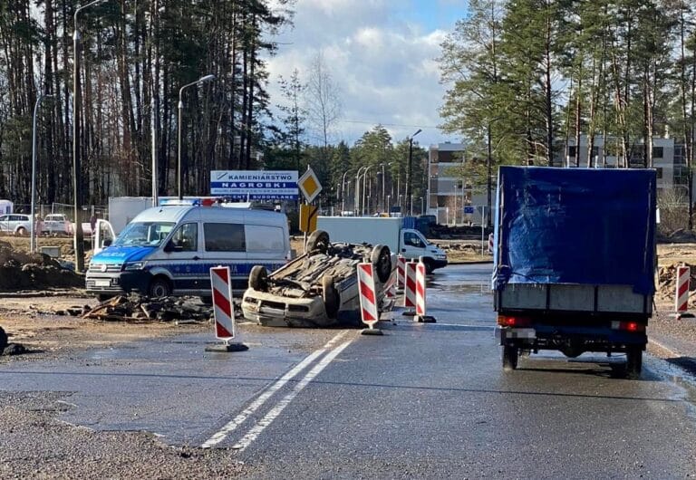 Rozbity samoch&oacute;d na drodze po dachowaniu, policja na miejscu zdarzenia, w tle drzewa i budynki.