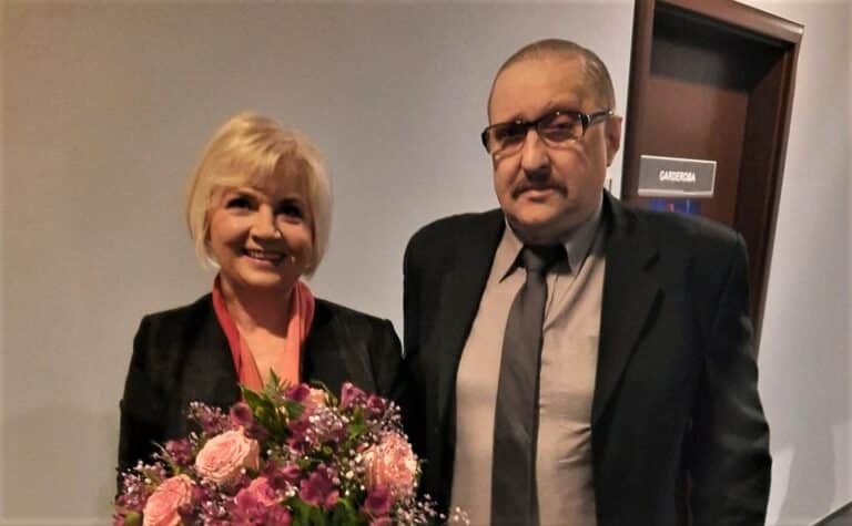 Dwoje ludzi na tle drzwi, kobieta trzyma bukiet kwiat&oacute;w, mężczyzna w garniturze.