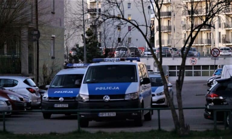Dwa policyjne radiowozy na parkingu w mieście, w tle bloki mieszkalne.