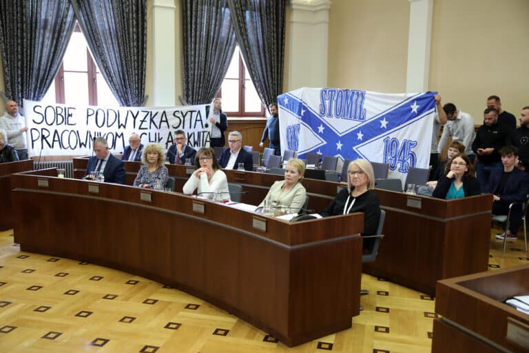 Radni podczas sesji rady miasta, za nimi transparenty i kibice Stomilu, w sali konferencyjnej.