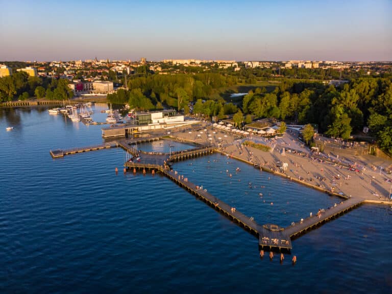Widok na plażę i marinę nad jeziorem Ukiel w Olsztynie, otoczoną zielenią i miastem w tle, podczas zachodu słońca.