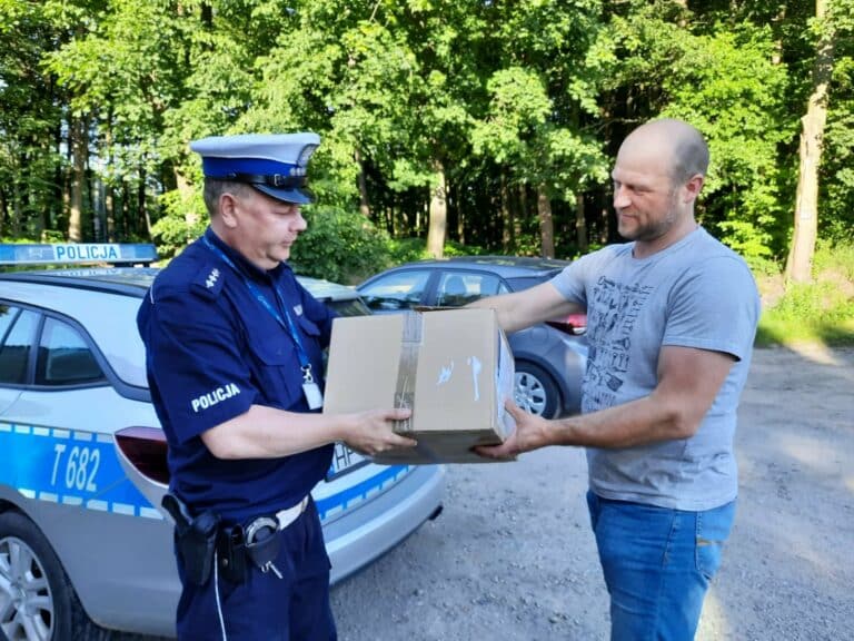 Policjant i mężczyzna wymieniają się paczką w pobliżu radiowozu na tle zielonych drzew.