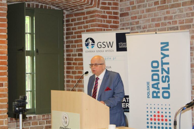 Mężczyzna w garniturze przemawia z mikrofonu podczas konferencji w pomieszczeniu z ceglaną ścianą i oknem.