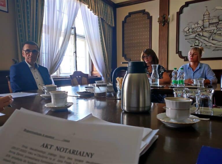 Spotkanie w eleganckim wnętrzu, na stole filiżanki, dzbanek i dokumenty, w tle okno z zasłonami.