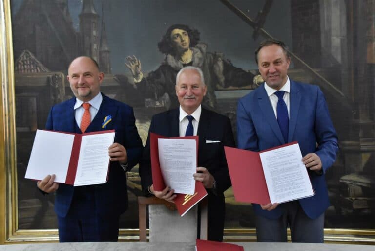 Trzech mężczyzn trzymających podpisane dokumenty podczas uroczystości w muzeum, w tle obraz Mikołaja Kopernika.