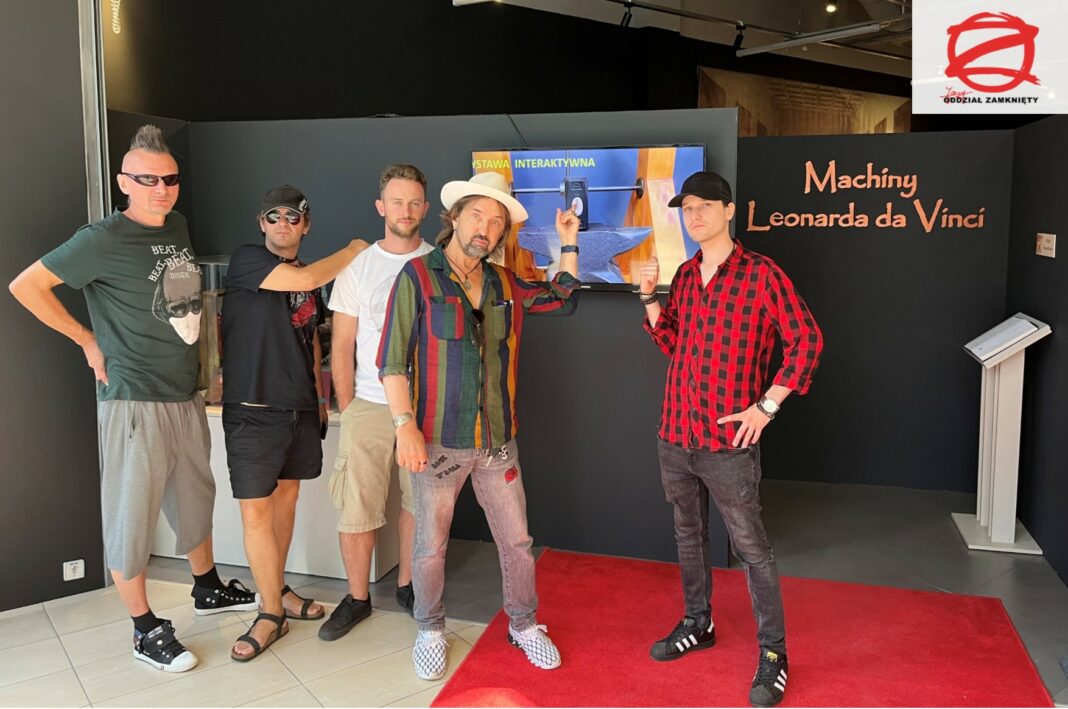 Grupa muzyków na wystawie o Leonardo da Vinci, stoją przed ekranem z napisem 'Machiny Leonarda da Vinci'
