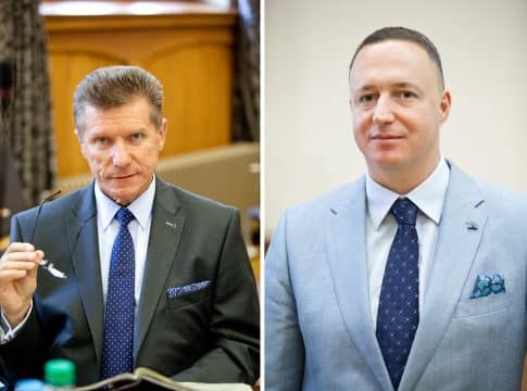 Zdjęcie dw&oacute;ch mężczyzn w garniturach, jeden z nich trzyma okulary, w tle pomieszczenie biurowe.