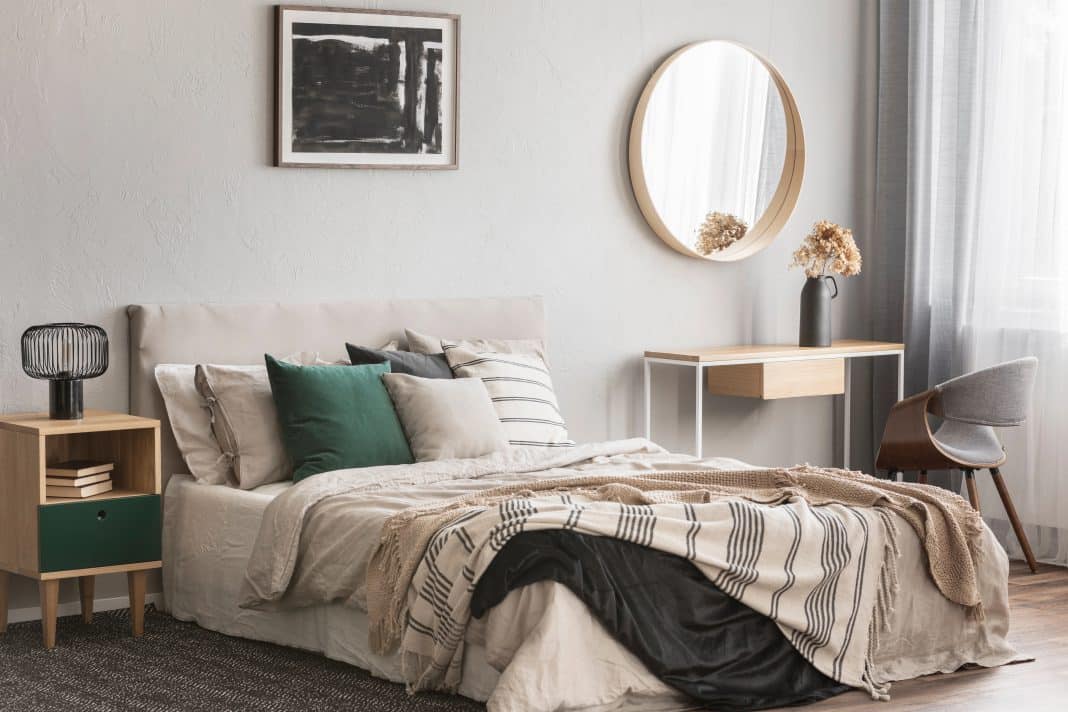 Elegant round mirror in wooden frame above fancy console table with flowers in vase in trendy bedroom interior with beige vase