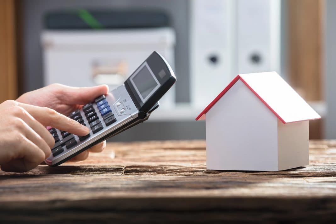 Human Hand Using Calculator With House Model On Wooden Desk