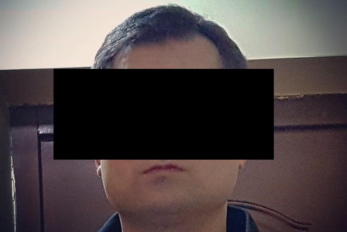 Zdjęcie mężczyzny z zasłoniętym oczami, ujęcie od pasa w górę, w tle ciemne drzwi.