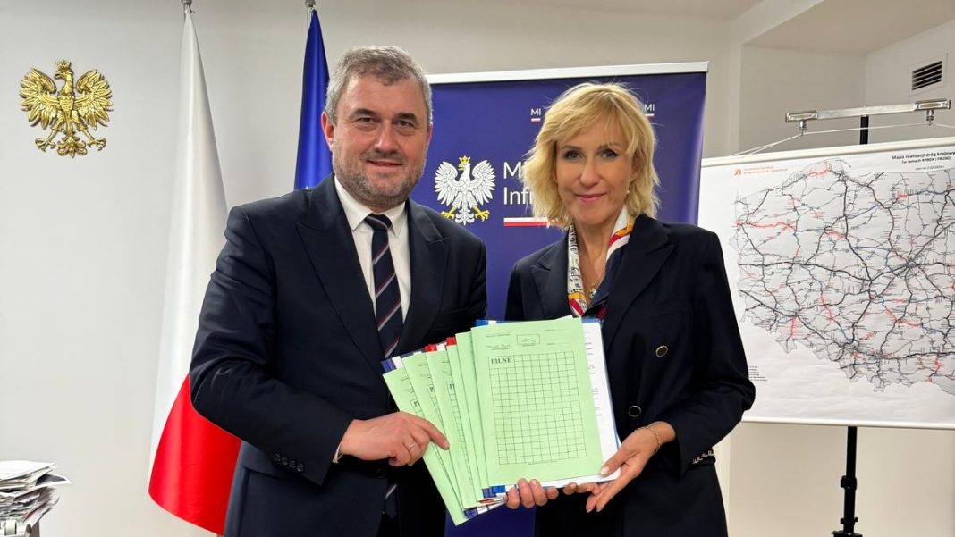 Dwoje os&oacute;b trzyma dokumenty podczas oficjalnej prezentacji inwestycji, w tle mapa i flagi.