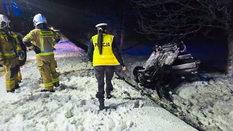 Samoch&oacute;d dachował na zaśnieżonej drodze, wok&oacute;ł strażacy i policjanci, nocne warunki.