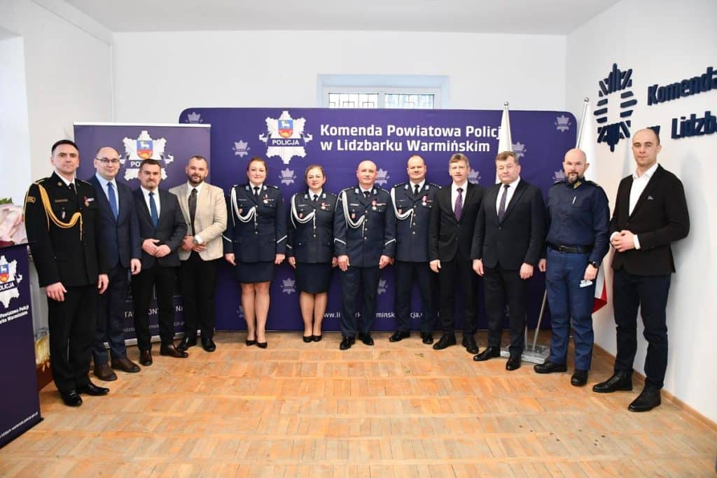 Grupa policjant&oacute;w i urzędnik&oacute;w w pomieszczeniu, za nimi tablica z napisem o komendzie w Lidzbarku Warmińskim.