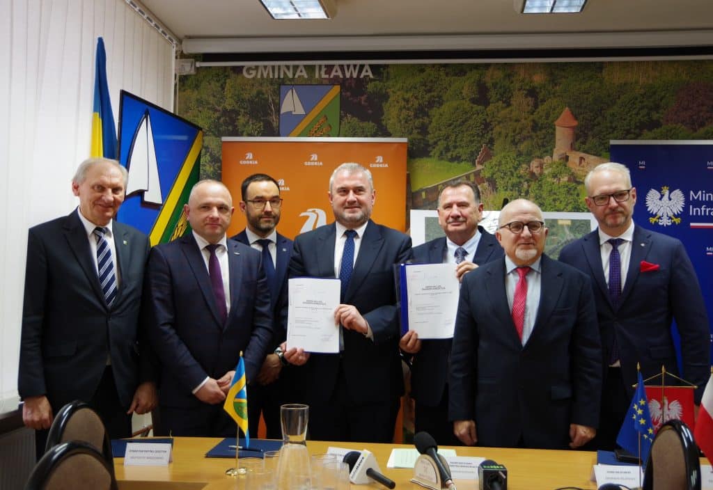 Grupa mężczyzn w formalnych ubraniach podczas podpisania umowy, za nimi logo gminy i plakat inwestycji.