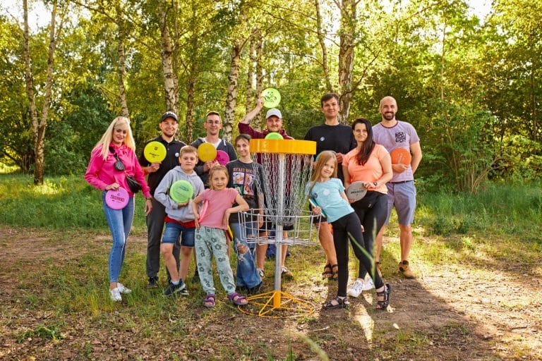 Grupa ludzi z disc golfami na tle lasu, dzieci i dorośli, podczas zimowych trening&oacute;w w Olsztynie.