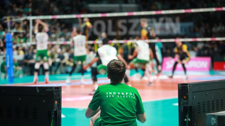 Młody kibic AZS Olsztyn obserwuje mecz siatk&oacute;wki w hali Azoty, w tle trwają emocje na boisku.